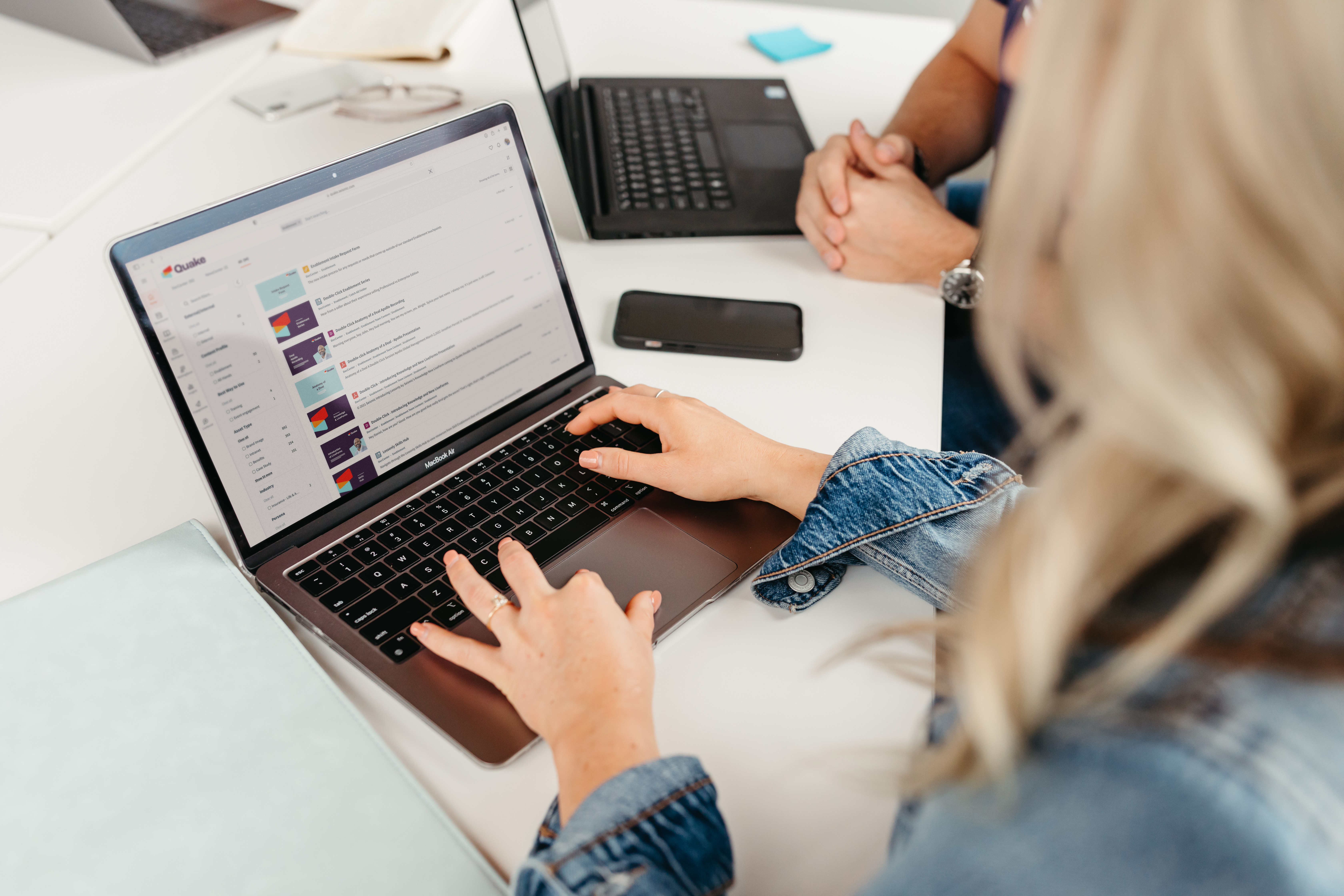 A person in a denim jacket uses a laptop on a desk alongside another individual and a smartphone.