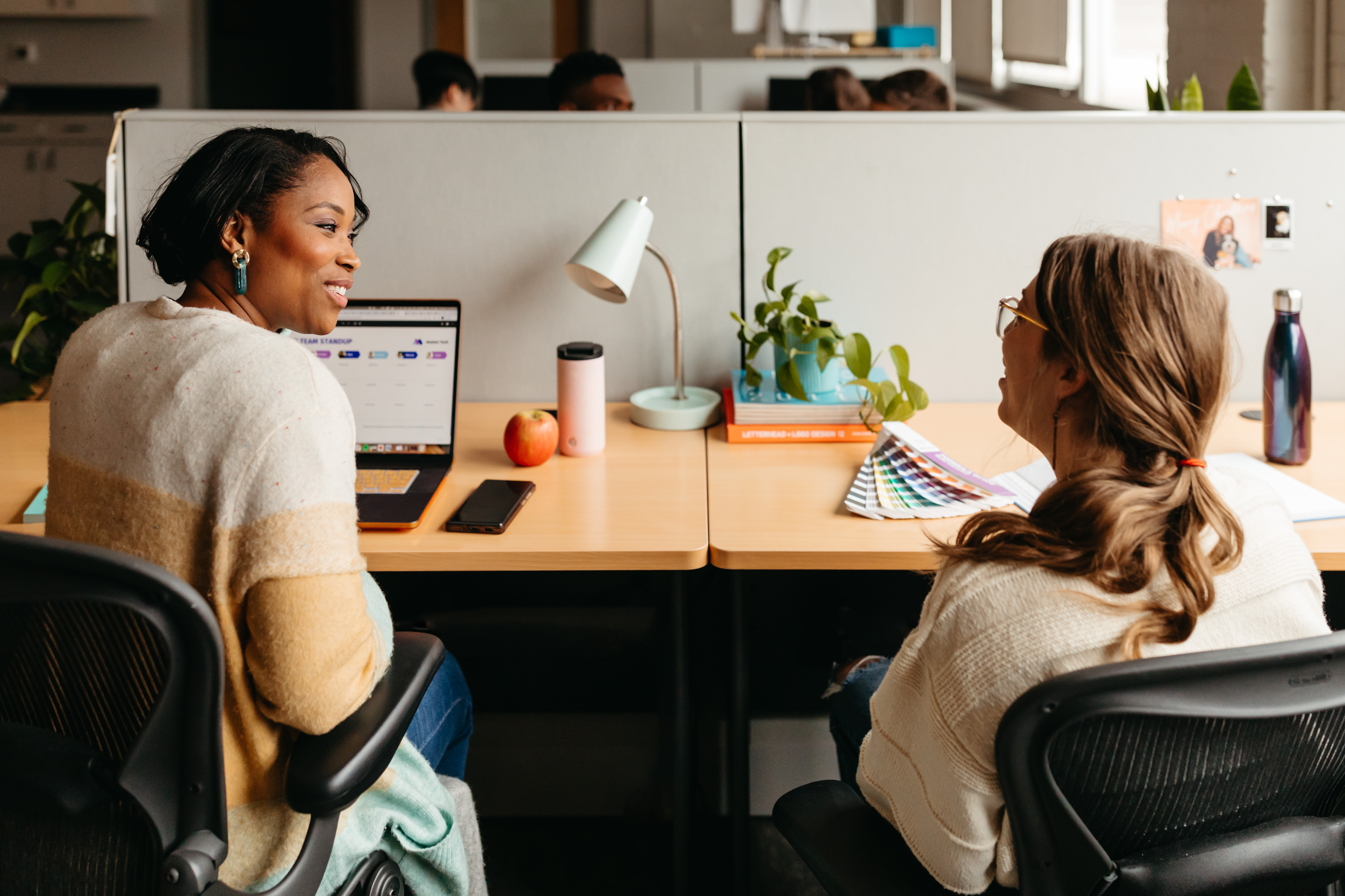 Two colleagues conversing at adjacent desks in a brightly lit office setting.
