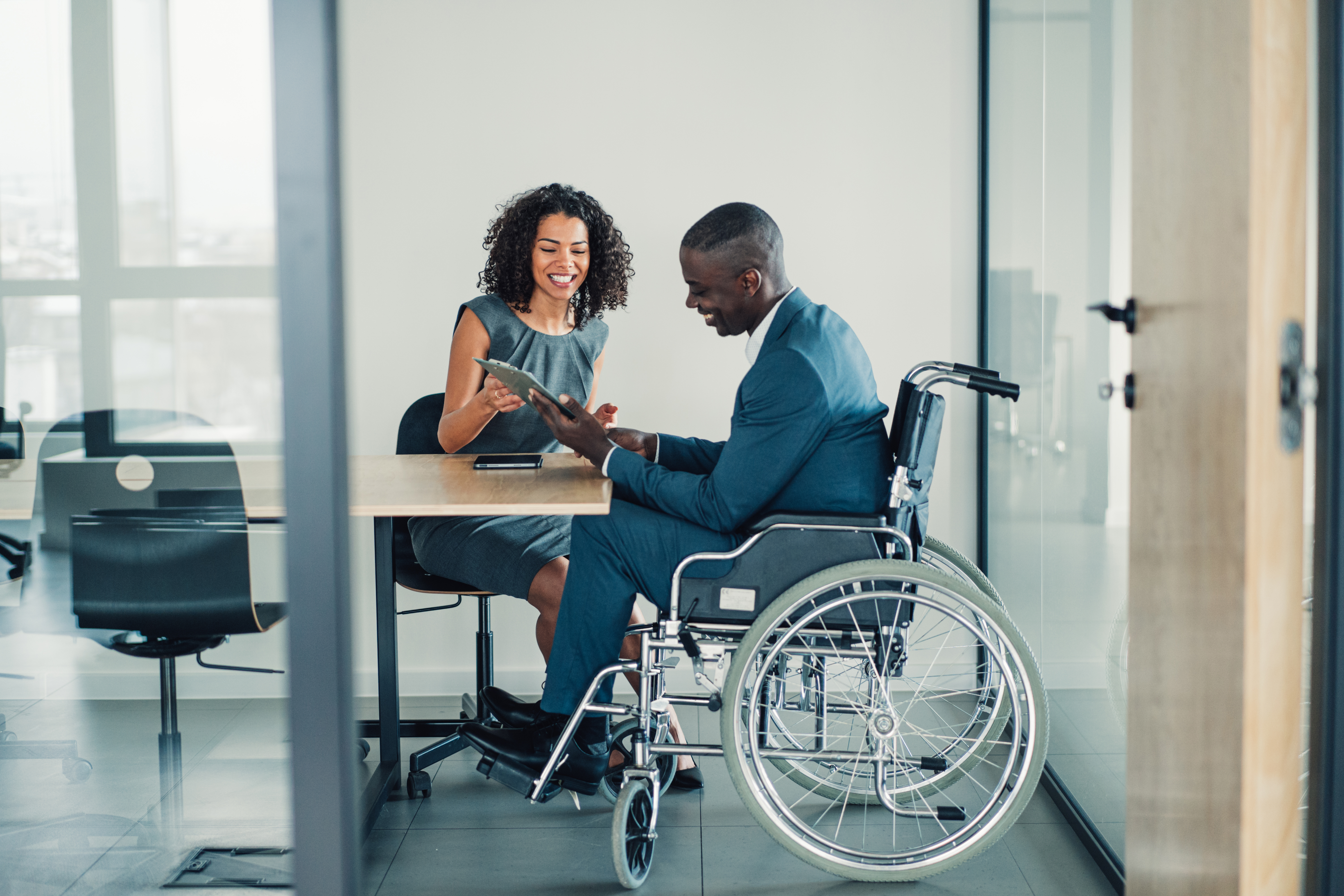 Two professionals in an office setting, one in a suit seated in a wheelchair holding a tablet and the other smiling in conversation.
