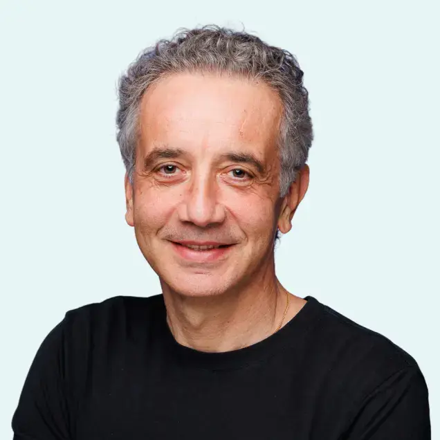 A person with short curly hair smiling, wearing a black shirt against a light blue background.